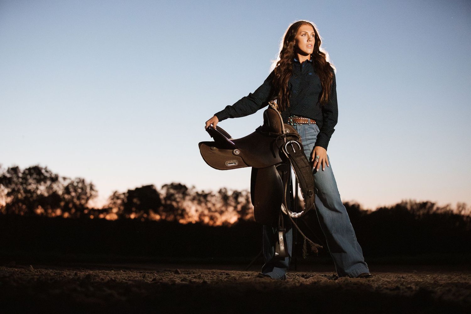 Casual outdoor portrait at dusk wearing black sweater and jeans against silhouetted trees.