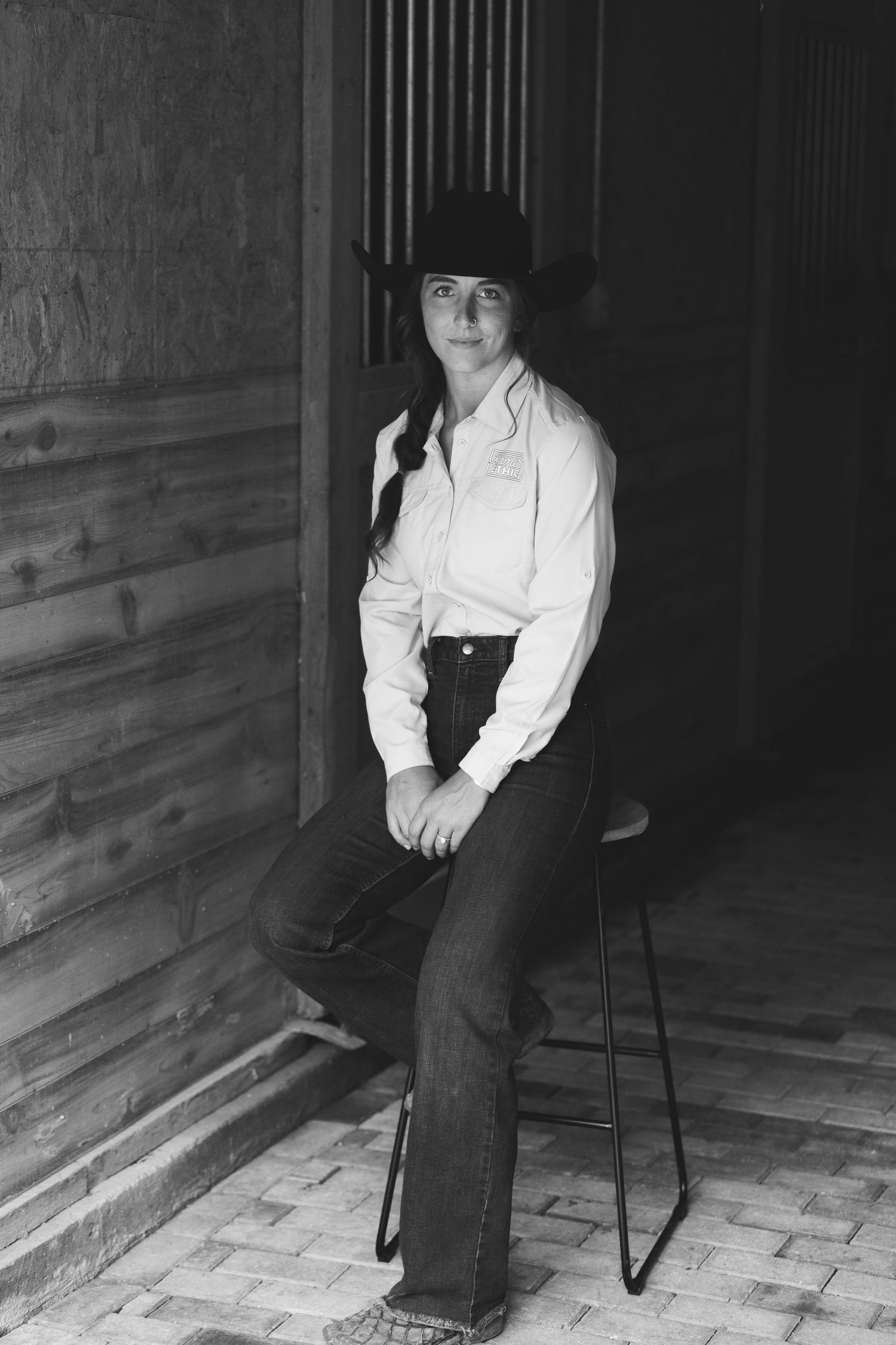 Woman in cowboy hat, white shirt, and jeans sits on stool near rustic wooden wall in black and white photo.