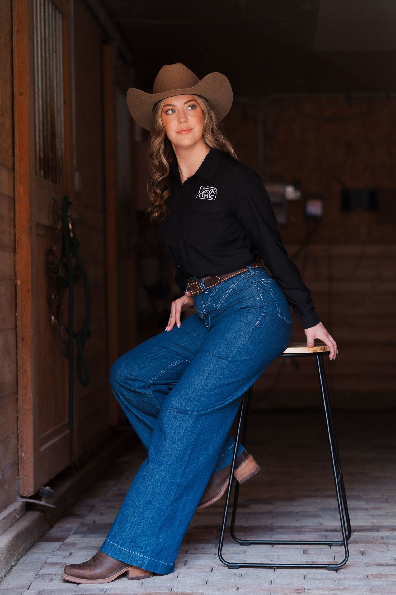 Woman in cowboy hat, black shirt, wide-leg jeans, and brown boots sitting on a stool in a rustic barn doorway.