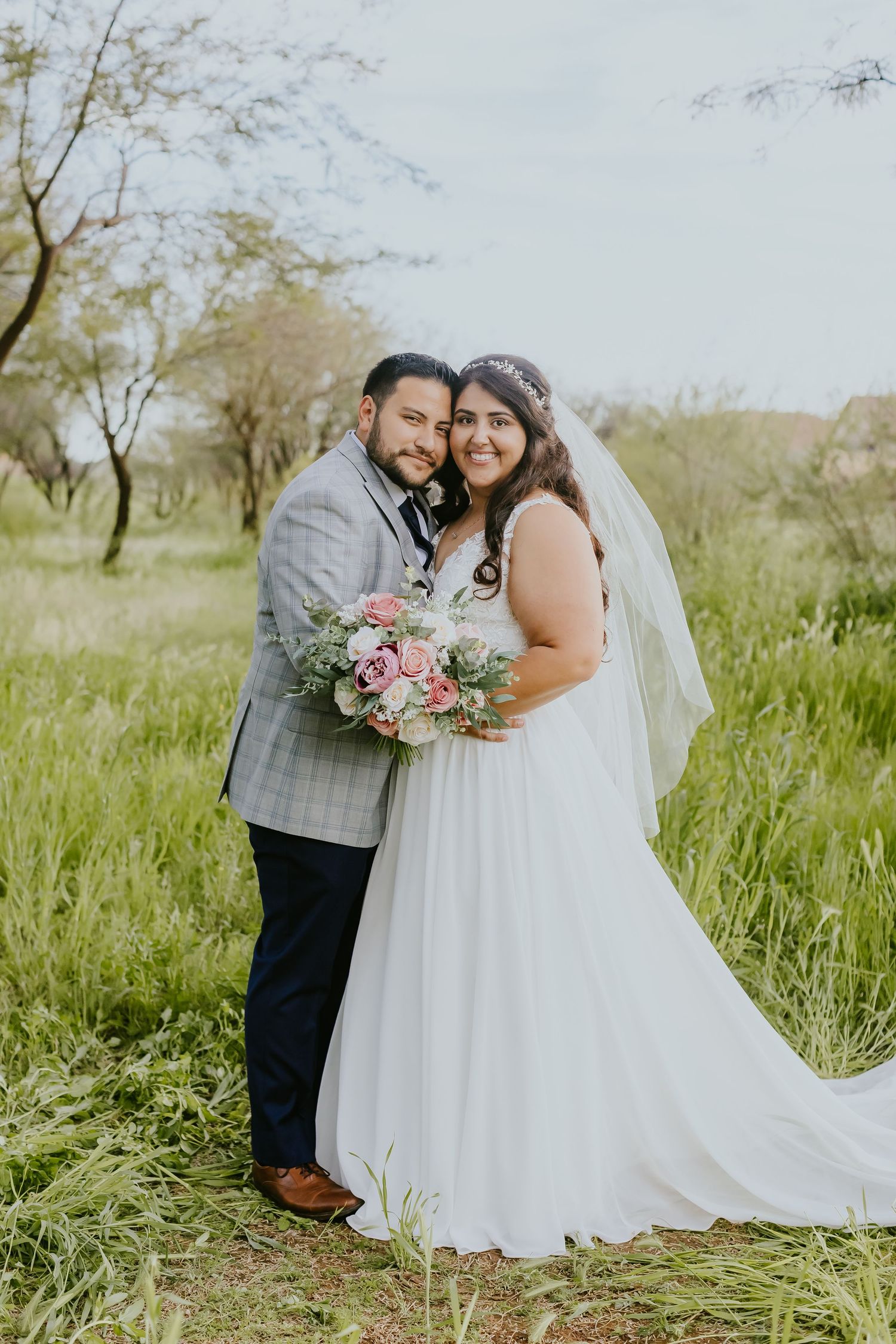 Wedding couple poses in meadow with pink and white bouquet and grey plaid suit.