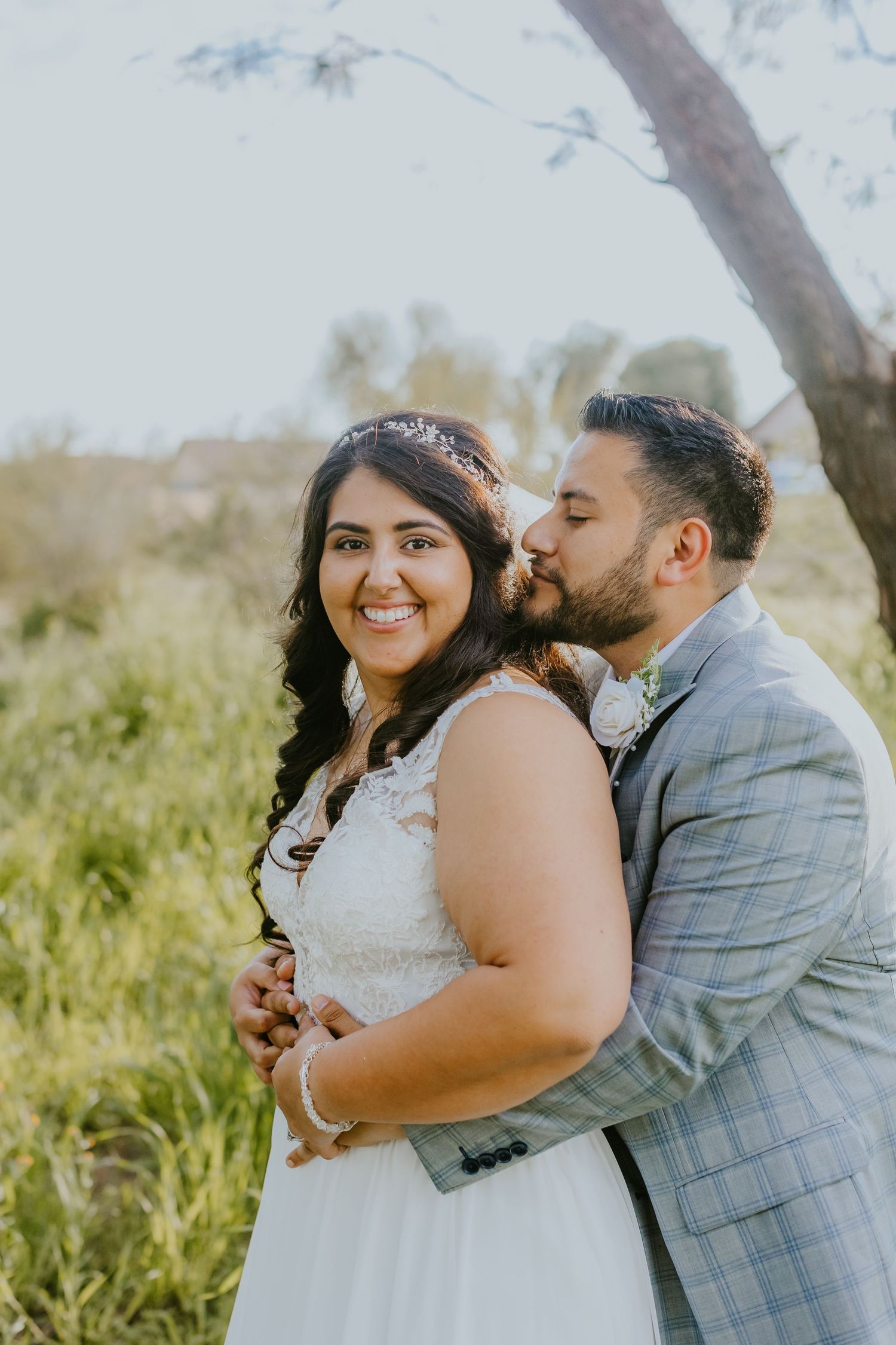 A romantic wedding photo of a couple embracing outdoors with the bride wearing a white dress and sparkly headpiece.