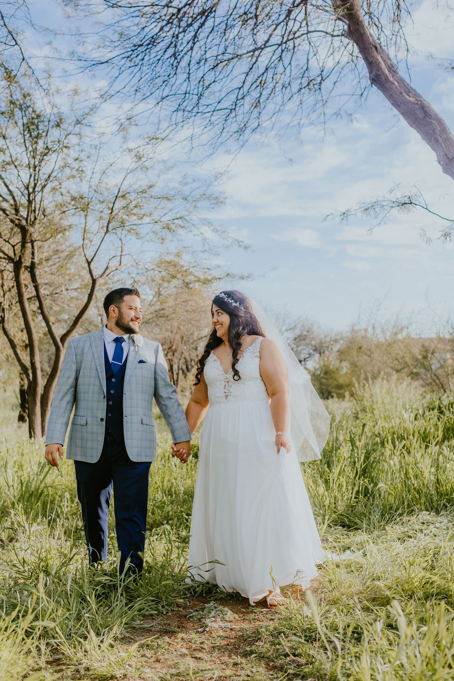 A wedding couple walks hand in hand through tall grass with trees in the background.
