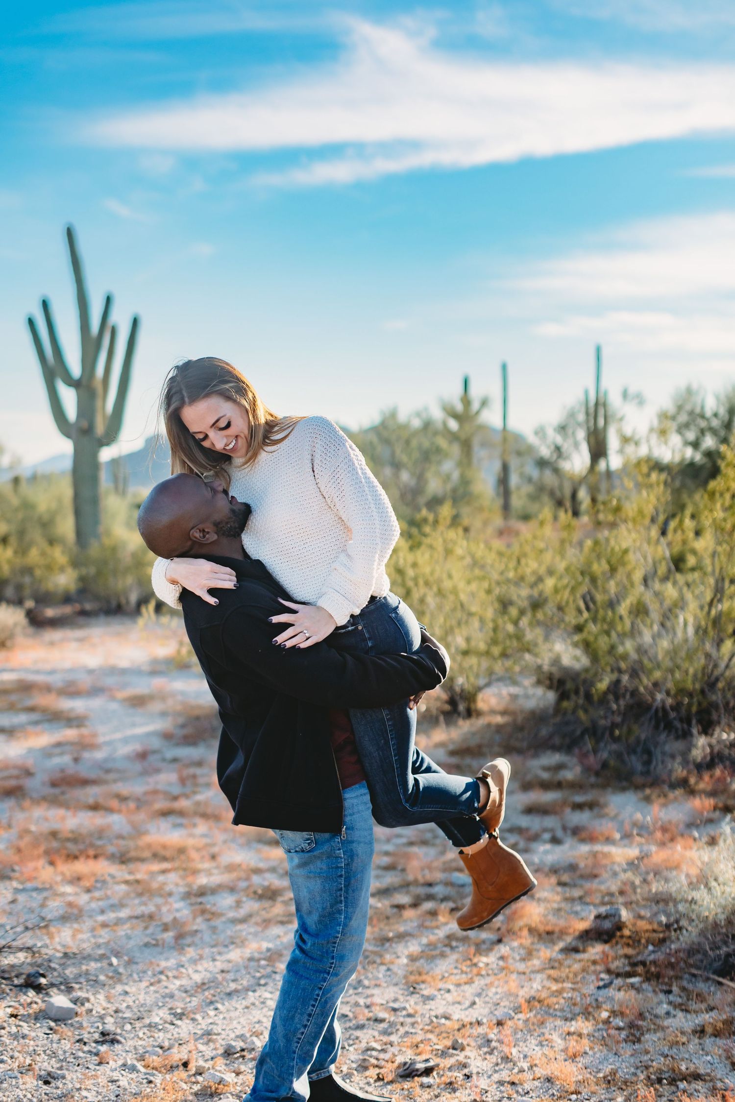 Playful couple embracing in a desert landscape with saguaro cacti and blue sky.