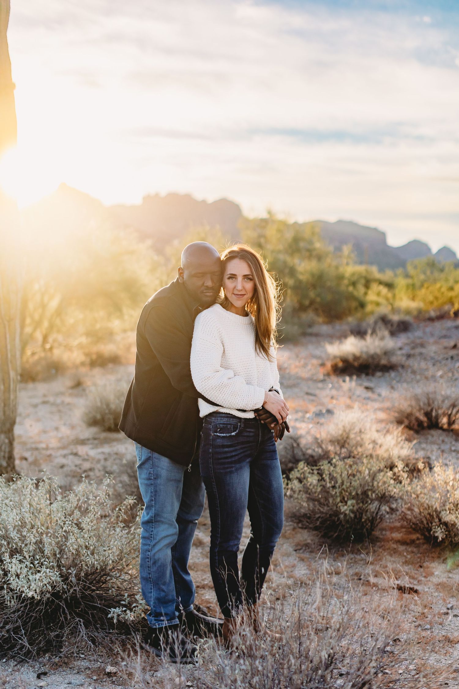 Couple embracing in desert landscape during golden hour with mountains in background.