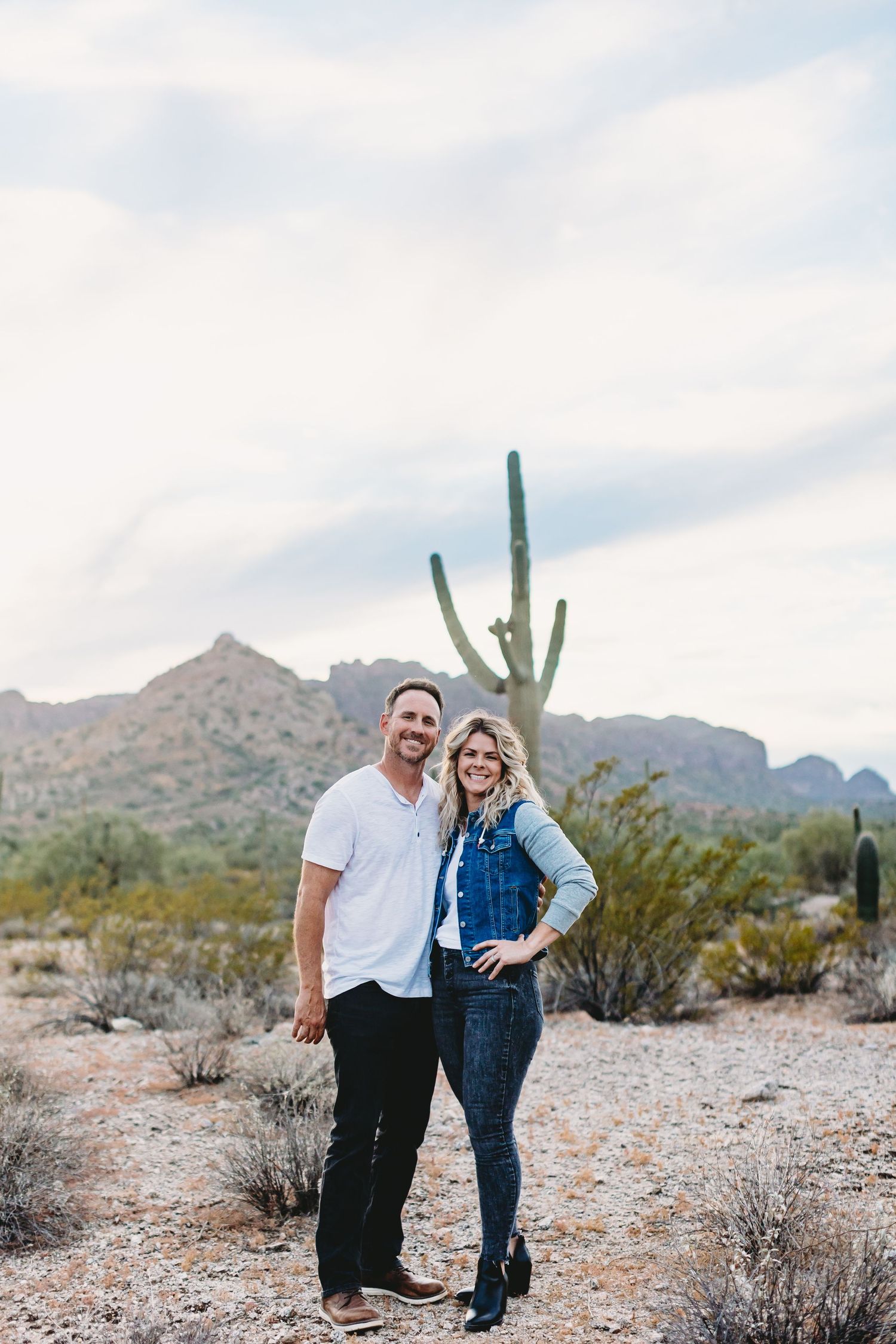 Casual couple posing in front of desert landscape with saguaro cactus and mountains.