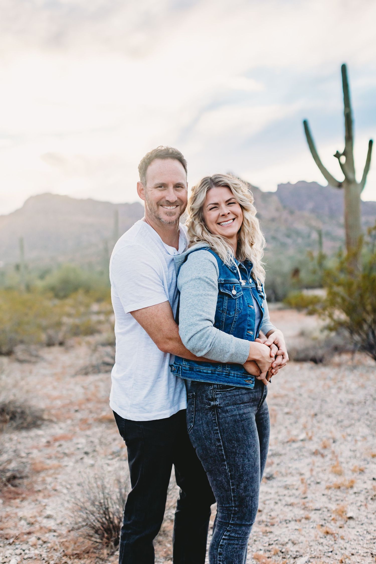 A couple shares a playful moment in the desert with saguaro cacti and mountains in the background.