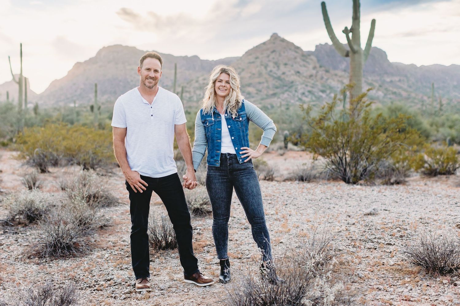 A couple embraces while standing on a desert trail surrounded by mountains and cacti.