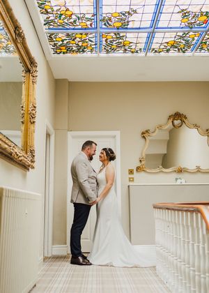 Stained glass skylight illuminates elegant interior staircase with ornate gold mirrors and white railings in historic venue.