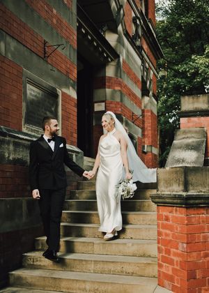 Bride and groom holding hands walking down brick building steps, bride in white halter gown with veil, groom in black tuxedo.