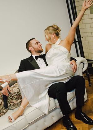 Joyful bride sitting on groom's lap on white sofa, both laughing, bride in white halter gown, groom in black tuxedo.