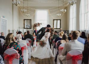An intimate indoor wedding ceremony with white chairs adorned in red sashes as guests watch the couple's romantic dip kiss.
