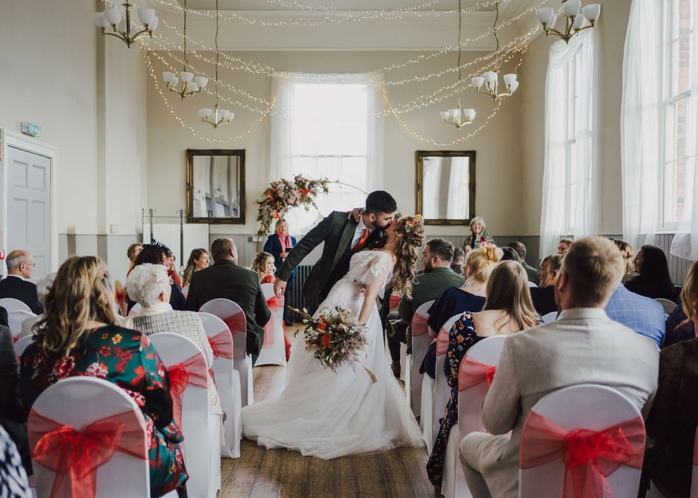 An intimate indoor wedding ceremony with white chairs adorned in red sashes as guests watch the couple's romantic dip kiss.