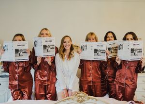 A group wearing matching burgundy jumpsuits holds up newspapers while one person stands in white in the middle.