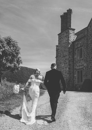 Black and white photo of a bride and groom walking hand in hand toward a stone manor house on their wedding day.