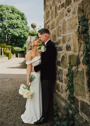 Bride and groom share a romantic moment against a stone wall, she holds white bouquet in off-shoulder gown, he wears black tuxedo.