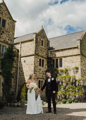 A bride and groom hold hands outside a stunning stone manor house adorned with wisteria on their wedding day.