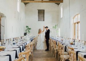 Bride and groom dancing in elegant barn wedding venue with long tables, white walls, and candlelit decor.