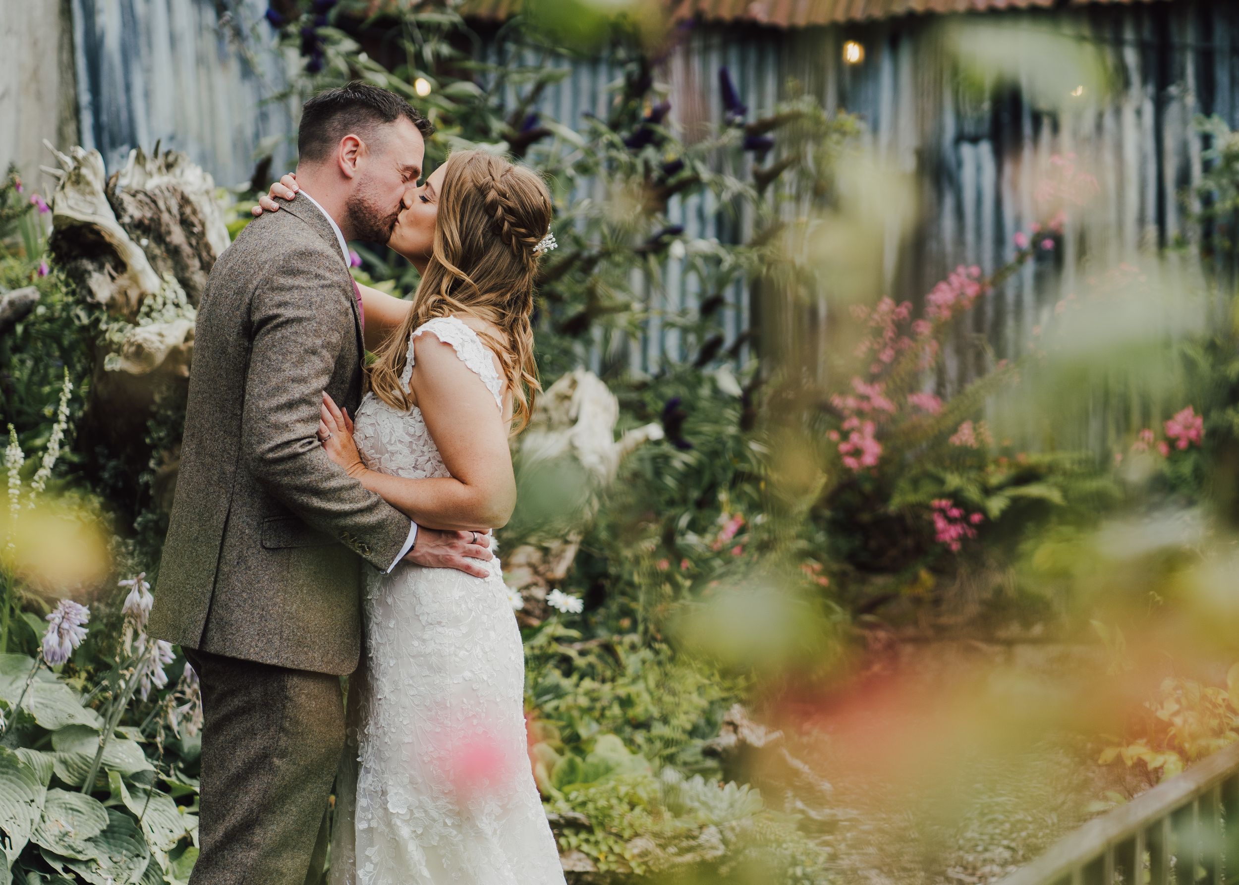 Intimate wedding portrait through garden foliage with soft focus flowers.