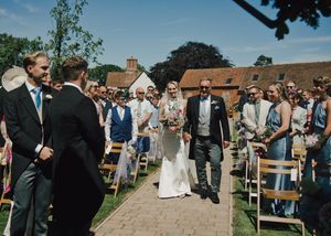 Bride walks down outdoor aisle with escort at sunny garden wedding, guests seated on wooden chairs.