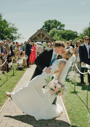 Bride and groom share a romantic dip kiss at outdoor wedding ceremony aisle, guests watching, sunny day.