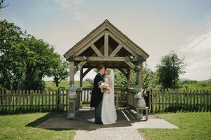 Bride and groom share a romantic kiss under a wooden gazebo in a lush green garden on their wedding day.