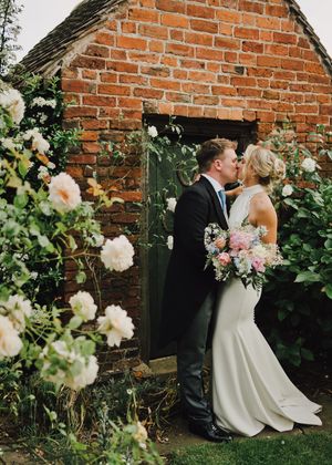 A bride and groom share a kiss by a rustic brick wall adorned with blooming roses, bride holding a pastel bouquet.