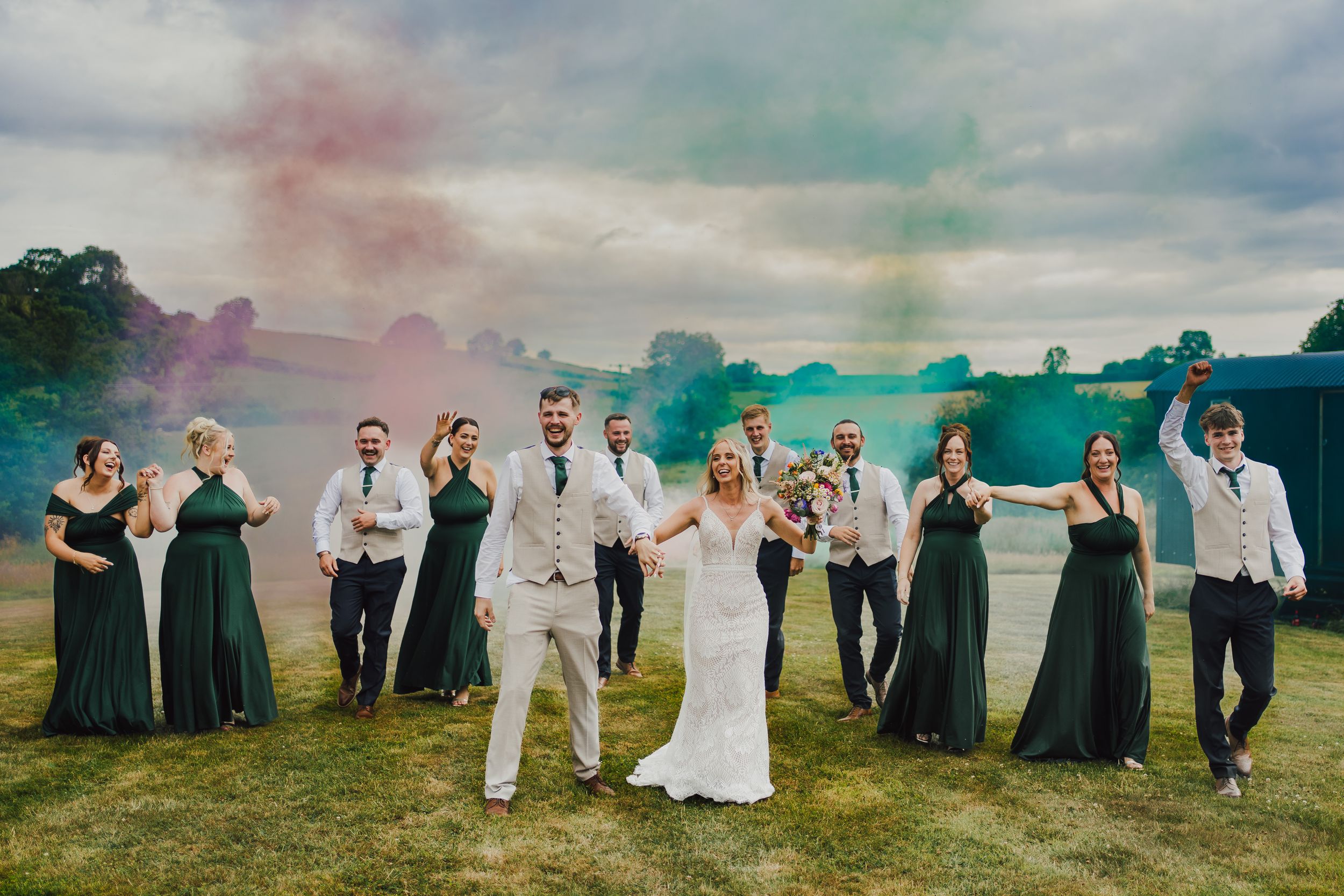 Wedding party dressed in formal attire poses in silhouette against dramatic cloudy sky on rolling green hillside.