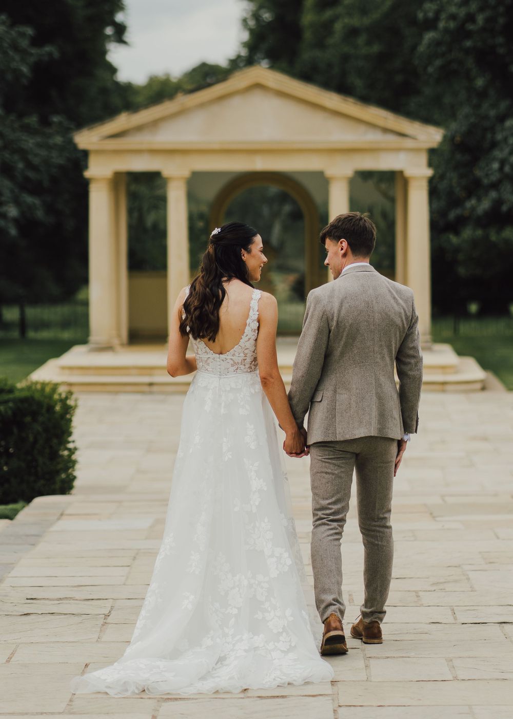 Couple walks hand in hand toward classical gazebo on stone pathway.