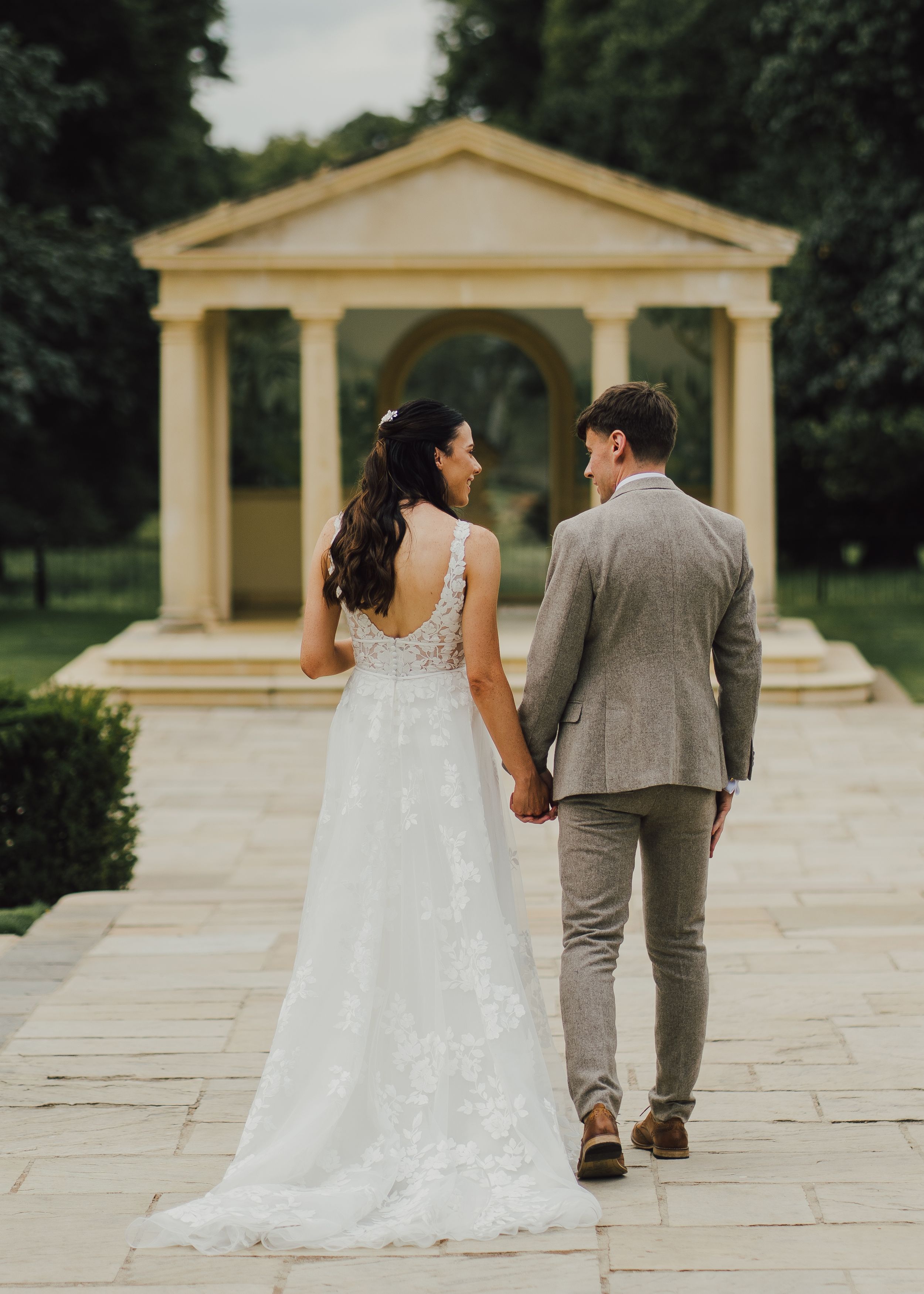 Couple walks hand in hand toward classical gazebo on stone pathway.