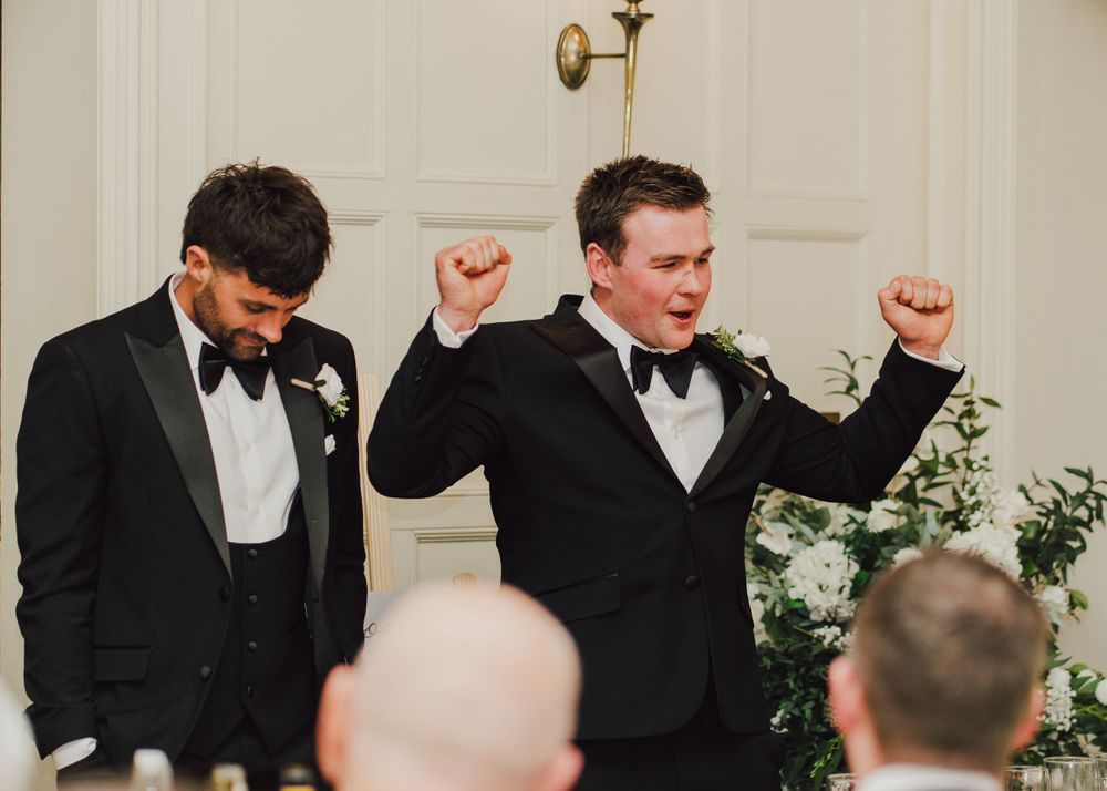 Two groomsmen in black tuxedos and bow ties celebrate joyfully during a wedding reception.