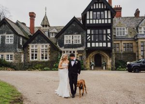 A couple and their brown dog stand outside a grand Tudor-style mansion with timber-framed architecture and historic details.
