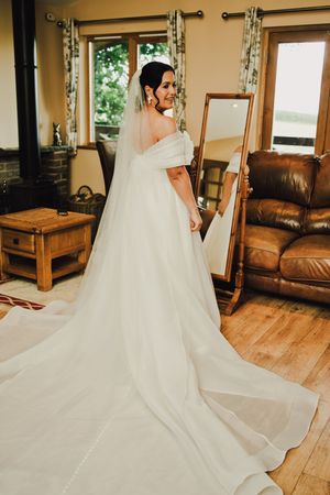 Bride in elegant white off-shoulder wedding gown with long train and veil, standing before a mirror in a cozy room.