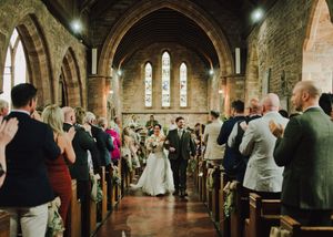 Bride and groom walk down the aisle of a historic stone church surrounded by applauding wedding guests.