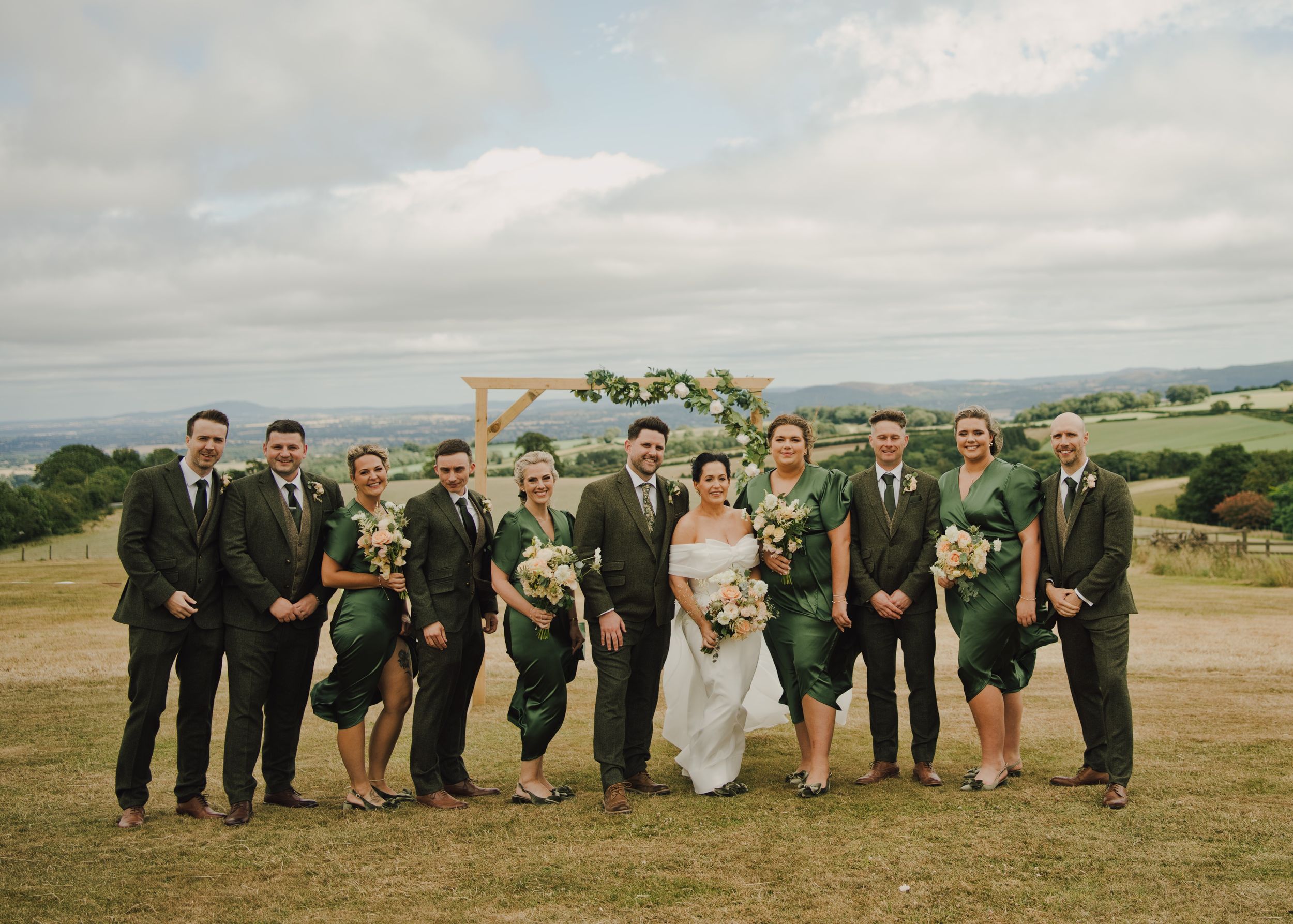 Wedding party in green attire poses outdoors on a hillside with a floral arch backdrop on a cloudy day.