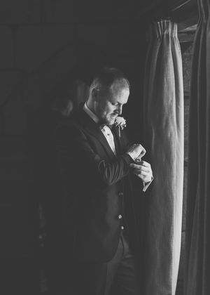 Groom in dark suit adjusting boutonniere by window in black and white wedding photo.