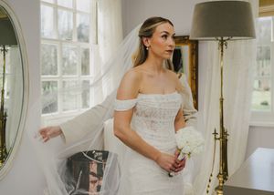 Bride in off-shoulder lace wedding dress holding white bouquet, wearing veil, standing by window in elegant room.
