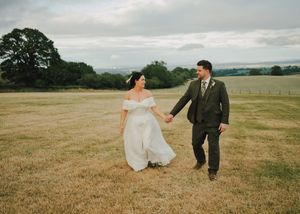Bride and groom holding hands walking through a golden field at their outdoor countryside wedding ceremony.