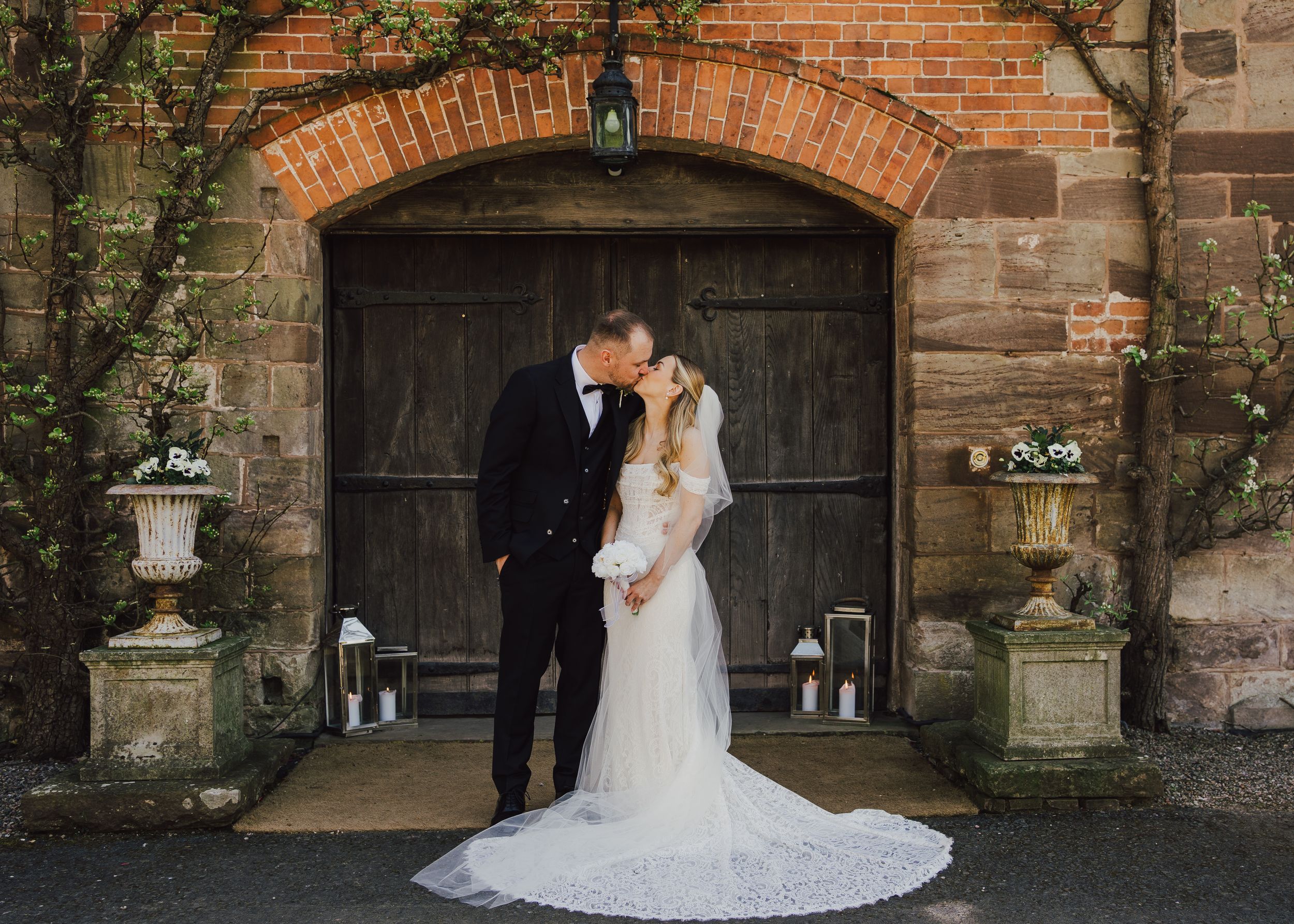 Bride and groom share a romantic kiss in front of rustic wooden doors at a brick wedding venue.