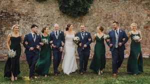 Wedding party in navy suits and emerald green dresses pose together against rustic brick wall outdoors.