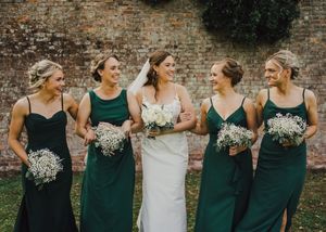 A bridal party in emerald green dresses with white floral bouquets stands together against a rustic brick wall.