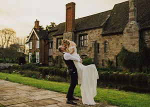 Groom lifting bride in white dress outside rustic stone English manor wedding venue.