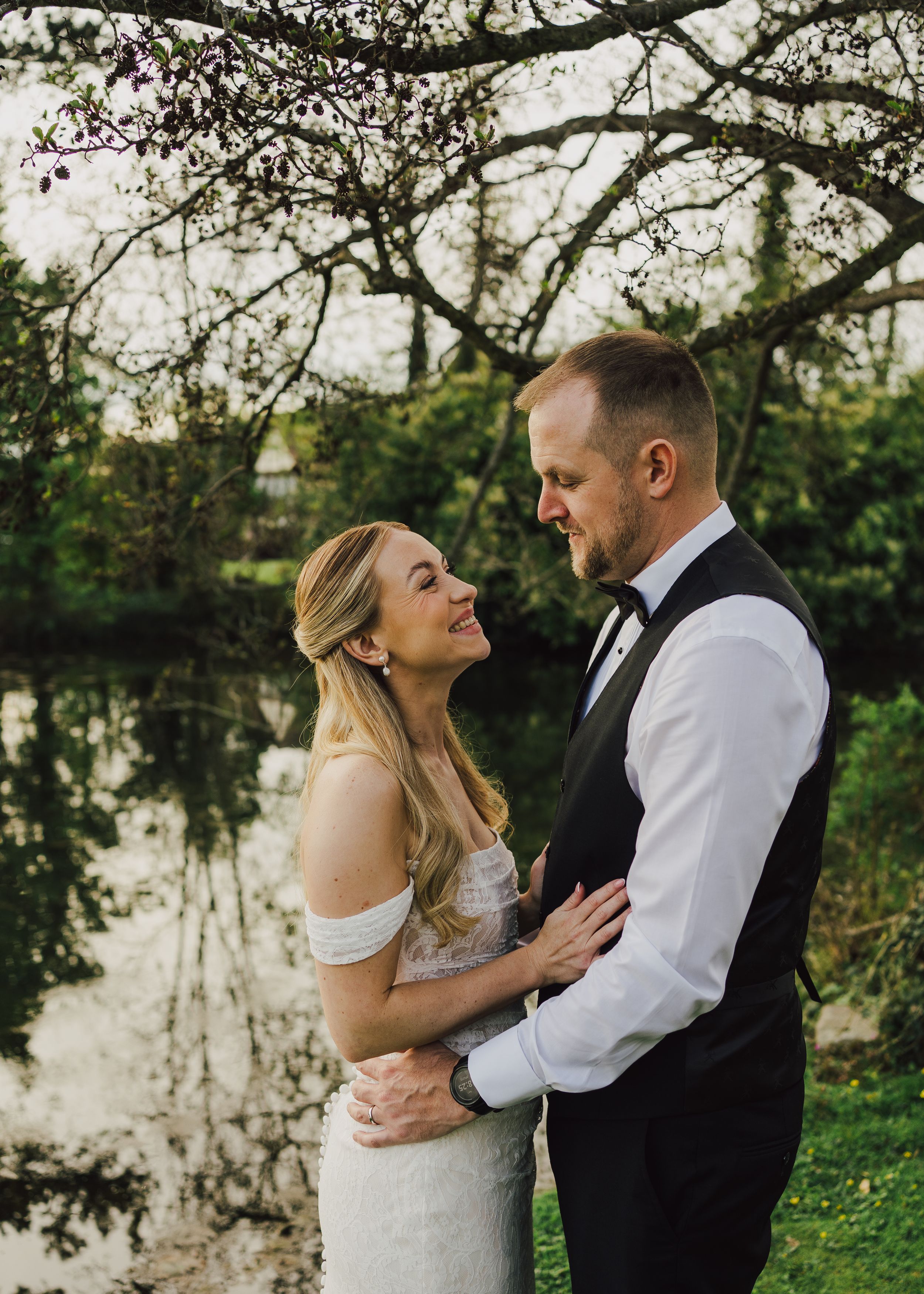 Bride and groom embrace romantically by a serene pond surrounded by lush greenery.