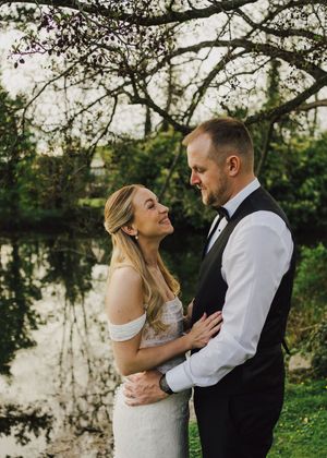 Bride and groom embrace romantically by a serene pond surrounded by lush greenery.