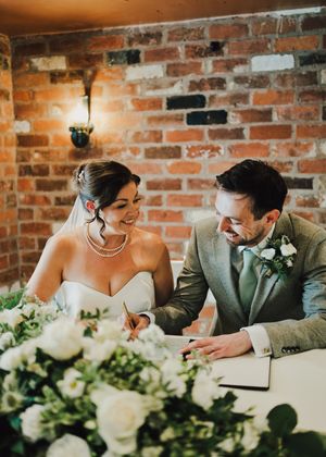 Bride and groom signing wedding register at rustic brick wall venue, surrounded by white floral arrangements.