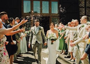 Newlywed couple walks through confetti shower surrounded by cheering guests outside brick building on wedding day.