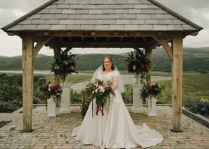 Bride in white gown holding bouquet under rustic wooden gazebo with floral arrangements and scenic hills backdrop.