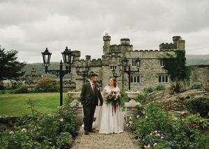 Bride and groom walking hand in hand in front of a stunning stone castle venue surrounded by lush gardens.