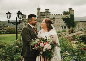 Bride and groom share a romantic moment in castle gardens, bride holding lush floral bouquet with pink and red blooms.