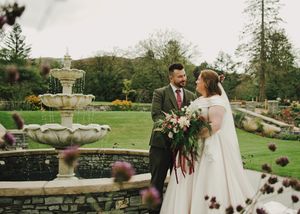 Bride and groom stand by stone fountain in lush garden, bride holding large floral bouquet in white gown.