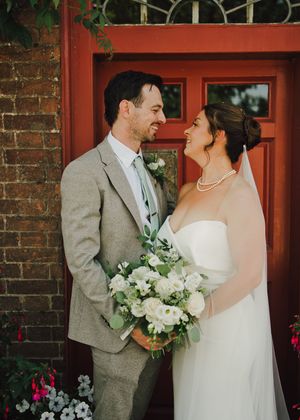 A bride and groom smile at each other in front of a red door, she holds a white bouquet and wears a veil and pearl necklace.